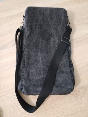 Vue de dessus d'une sacoche rectangulaire en denim gris foncé, de style upcyclé, posée à plat sur un plan de travail en bois clair. On la voit de face, au tiers supérieur on y voit un écusson rond marqué au fer chaud du logo de Basance. Une bandoulière noire réglable est fixée sur les côtés de la sacoche en haut par des anneaux métalliques en D.